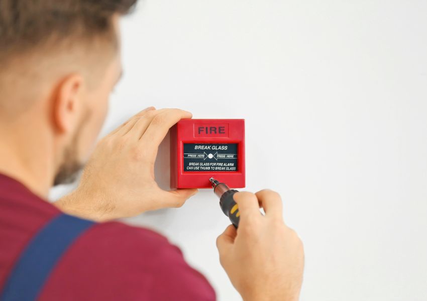 A property manager installs a red fire alarm in a landlord’s investment property with a screwdriver.